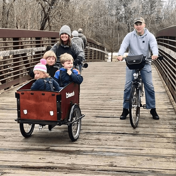 mom riding cargo bike with 3 kids over bridge next to biking dad  #color_Classic Sedona Woodgrain