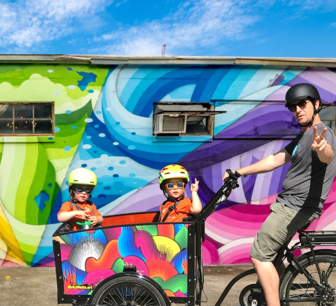 Ricky Watts on bunch bike with two kids in front of rainbow mural