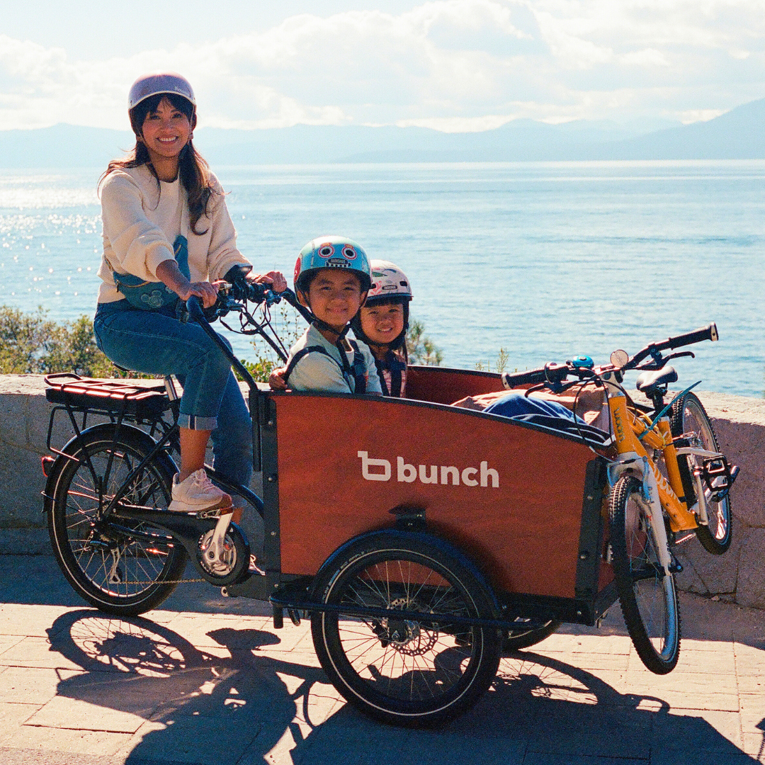 Two kids in a bunch bike with mom carrying a woom kids bike on front panel - @itsterryberry #color_Sedona Woodgrain