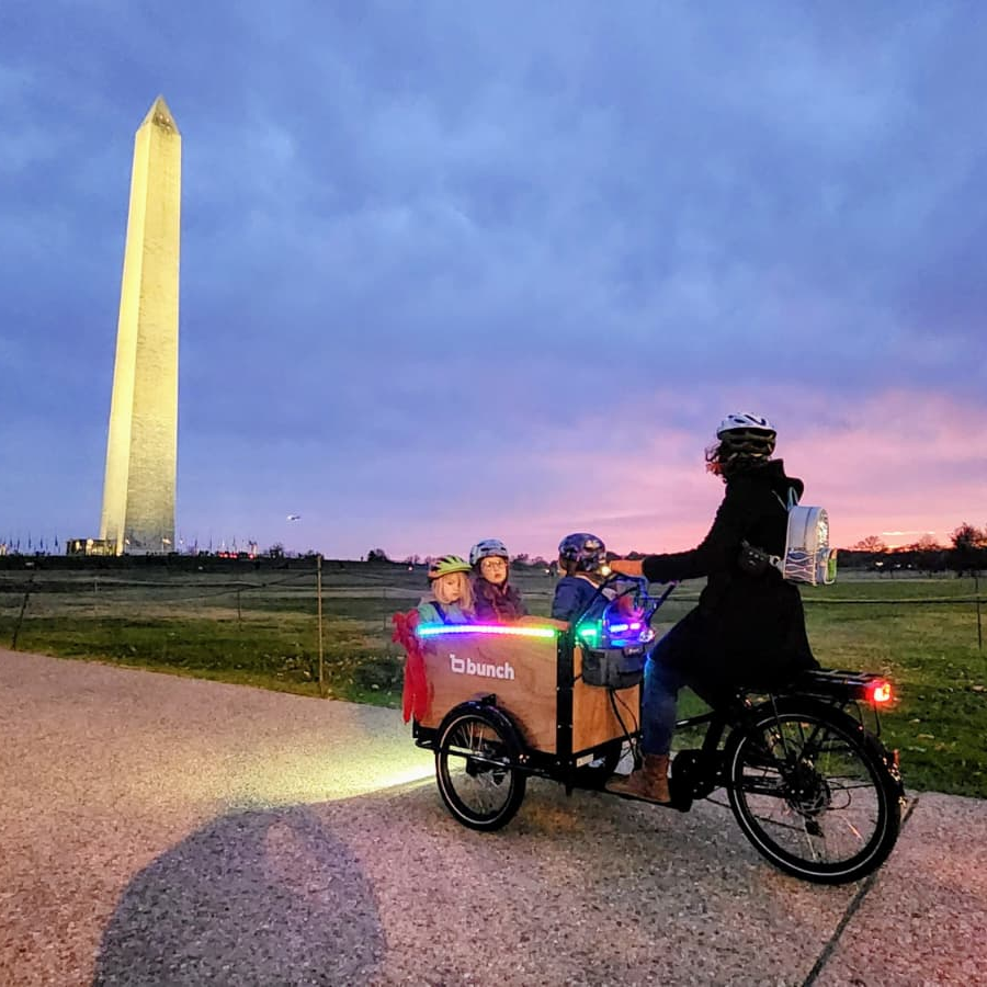Person riding a cargo bike with children at sunset near the Washington Monument. #color_Honey Woodgrain